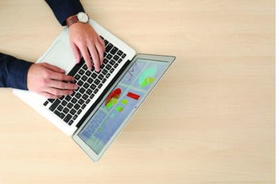 Hands typing on a laptop on a wooden table. The laptop screen is displaying charts.