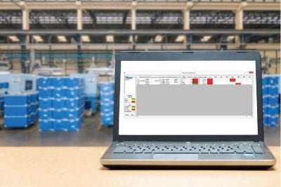 Laptop computer sitting on a wooden table in a warehouse. Blue bins are stacked up behind the monitor. Operation dashboard is displayed on the screen for shop floor data collection