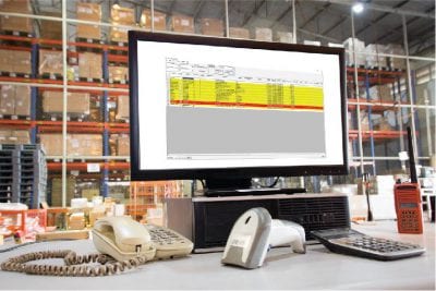 Computer sitting on a grey table. On the screen is a dashboard. In front of the computer is a barcode scanner, calculator and phone. Behind the computer are cardboard boxes and wooden pallets on shelves for shop floor data collection