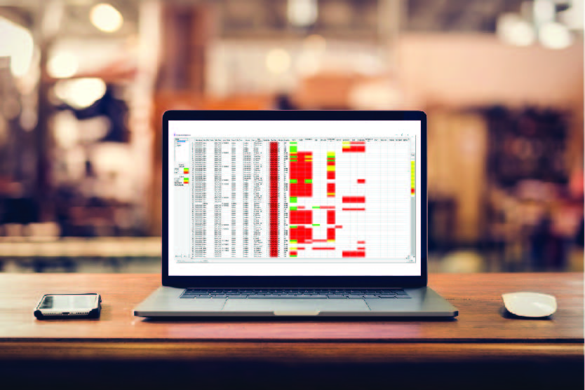 Laptop sitting on a wooden desk in an office with a phone and wireless mouse sitting beside it. On the screen is displayed scheduling dashboard for shop floor data collection