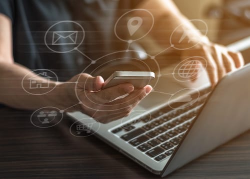 A man holding a cellphone while scrolling on a laptop with his other hand.