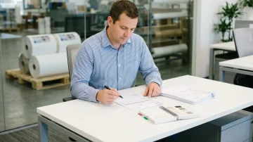 Manufacturing procurement specialist reviews a purchase order in a modern office with warehouse visibility, demonstrating efficient operations.